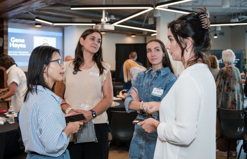 Four women actively conversing at a professional event with a presentation screen in the background.